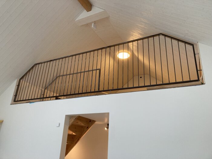 Svart räcke monterat längs kanten av en loftvåning i ett rum med vitmålat tak och väggar, belyst av en inbyggd taklampa.