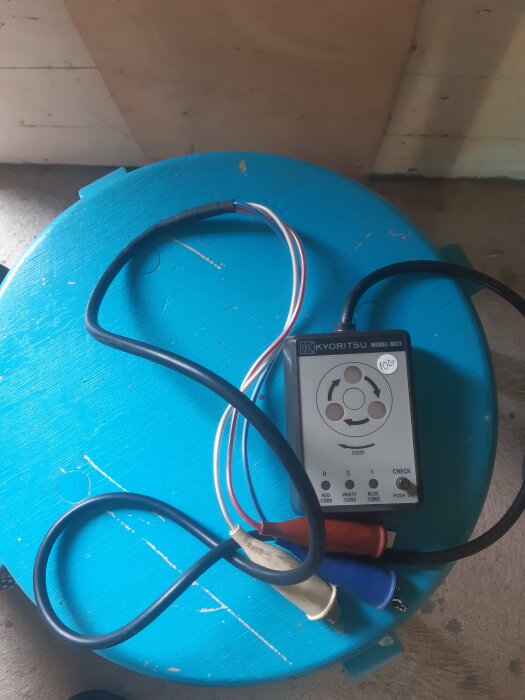 En Kyoritsu rotationsriktningsvisare med kablar på en blå yta, omgiven av tre testsladdar i rött, vitt och blått.