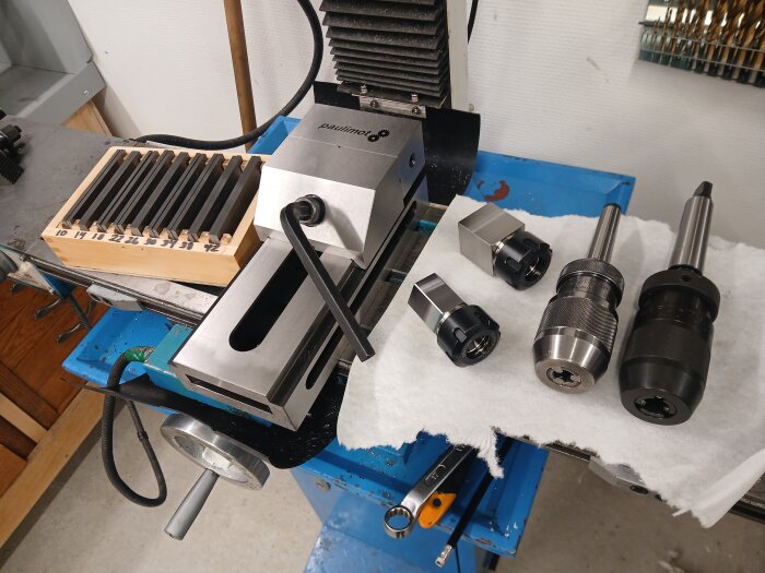 Fräsmaskin med verktyg och tillbehör, inklusive snabbchuckar och skruvstycke, samt uppmärkta verktyg i en trälåda på arbetsbänken.