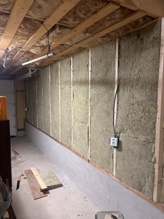 Isolering monterad på väggen i en garage- eller förrådslokal med träreglar, isoleringsskivor och en synlig elkontakt. Golv och isoleringstillbehör synliga.