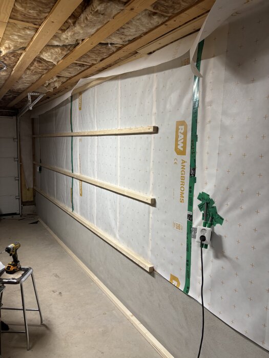 Vägg med ångbroms och isolering på träreglar, eluttag på väggen; verktyg på en stol i förgrunden, ofärdigt rum.