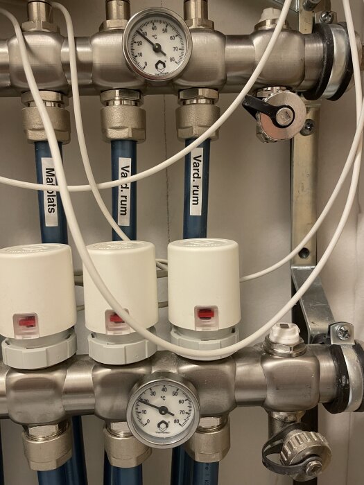 Golvvärmesystem med termostater och temperaturmätare i ett teknikrum med märkta rör för olika rum.