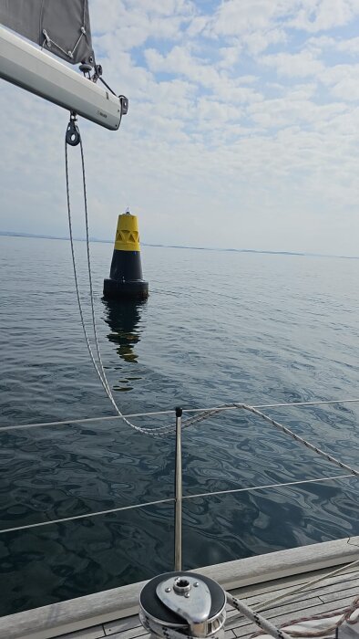 Segelbåt på lugnt vatten bredvid en gul och svart boj under en molnig himmel.