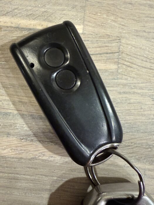 Svart fjärrkontroll med två knappar och fäste på en träribbad yta.