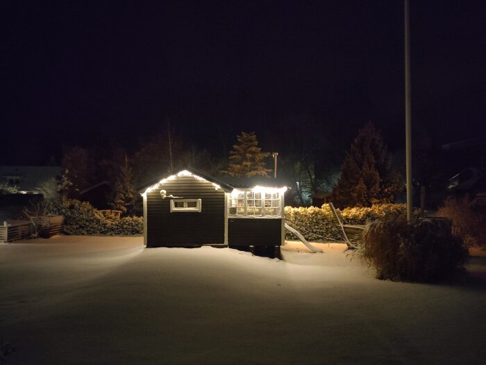 Belyst byggnad i en snötäckt trädgård på natten, omgiven av mörker och snö som lyser upp omgivningen.