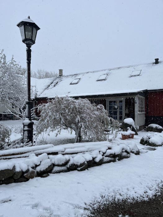 Snötäckt gård i Skåne med snö på ett staket av stenar. En lampa och ett snötäckt rött hus syns i bakgrunden.
