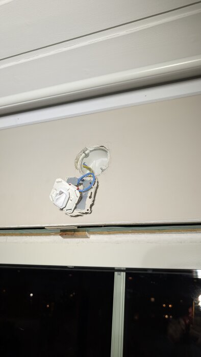 Vägg med synligt installationshål och löst sittande eluttag, kablar hänger ut. Under fönstret syns mörkt nattlandskap.