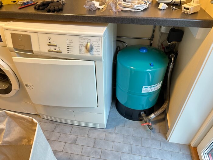 Blå hydrofor installerad under en bänkskiva i en trång tvättstuga, bredvid en vit torktumlare, med rör och ventiler anslutna.