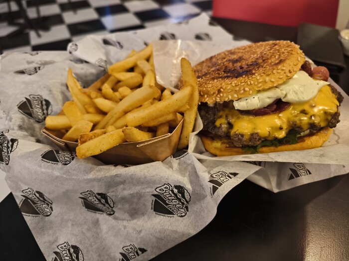 Cheeseburgare med vitlök och pommes frites på ett papper med Burger House-logotyp, serverade på en svart bordsyta.