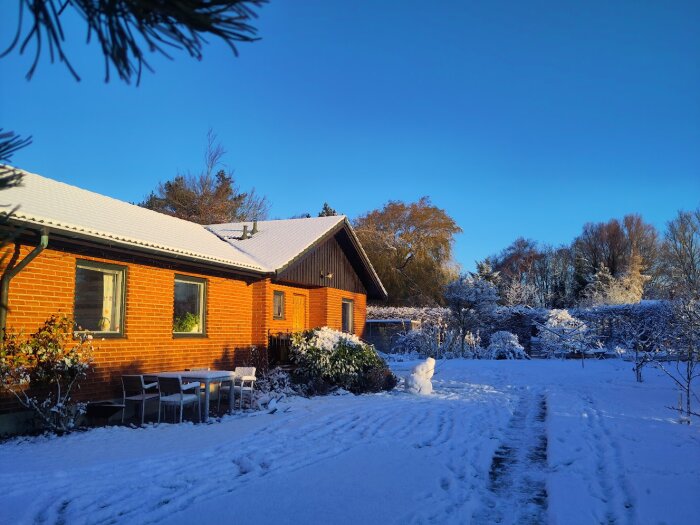 Snötäckt trädgård framför ett rött tegelhus med snö på taket och bortglömda trädgårdsmöbler i snön.
