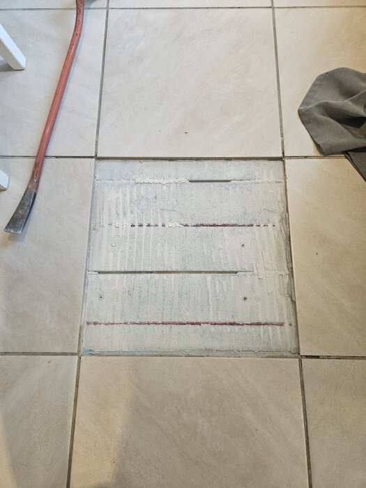 Öppnad golvplatta med synliga värmeslingor under kakel, bredvid ligger en röd kofot och en grå trasa.