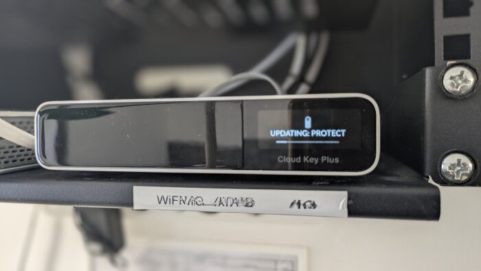 En Cloud Key Plus-enhet som uppdateras, visas på en hylla med texten "UPDATING: PROTECT" och etikett "WIFI RMC LADAMO" nedanför.