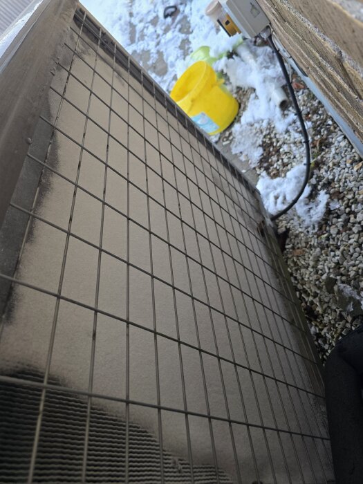 Baksidan av en pump med ett galler, sladd, och snö på marken, en gul hink syns i bakgrunden.