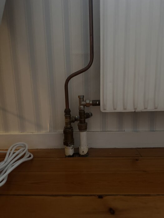 Radiator med kopplingsventiler och kopparrör anslutna, placerad mot en randig vägg och trägolv.