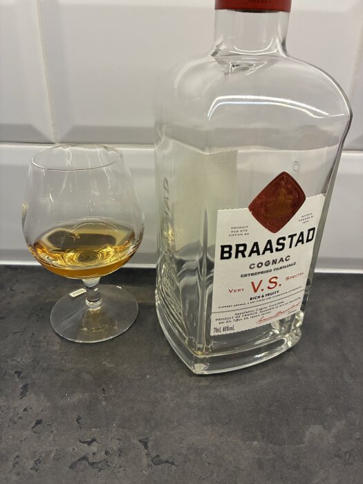 En flaska Braastad cognac och ett glas med cognac står på en bänkskiva framför en vit kakelvägg.