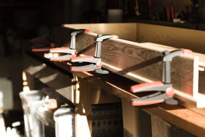 Träbitar fastklamrade med flera limklämmor på en arbetsbänk under byggandet av ett skrivbord av furu.