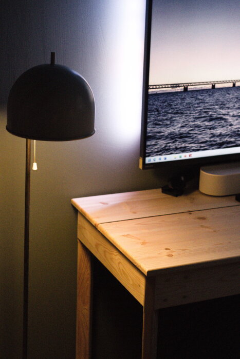 Furuskrivbord med frästa kanter, inspelat i ett rum med en lampa och en datorskärm som visar en bild på en bro över vatten.