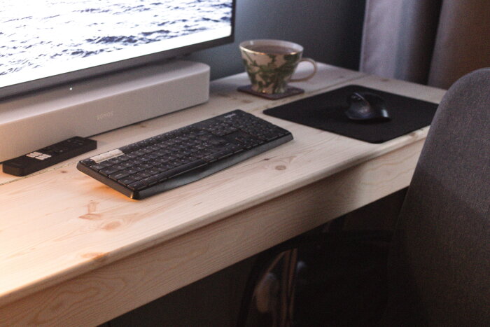 Hemmagjort skrivbord i ljust trä med datormusmatta, tangentbord och kopp; bildskärm synlig i bakgrunden.