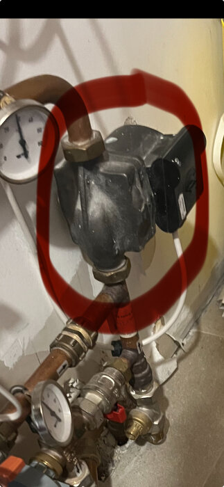 Gammal cirkulationspump med rör och tryckmätare i ett värmesystem, markerad med röd cirkel.