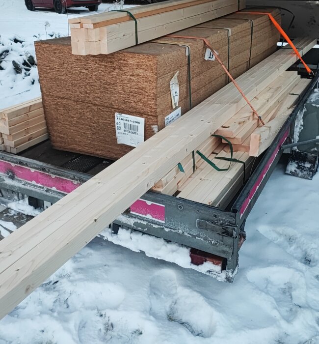 Material inklusive träbalkar och OSB-skivor ligger på ett släp i ett snöigt utomhusområde.