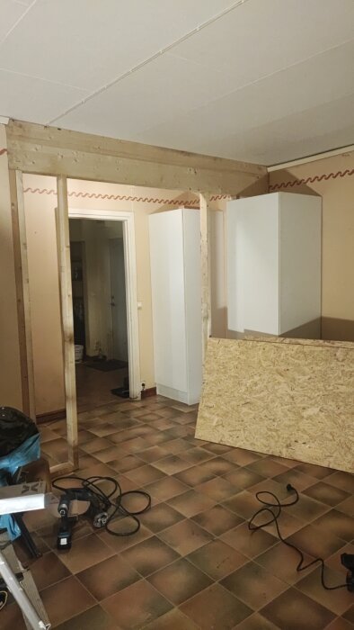 Renovering påbörjad i rum med träbalk på plats, två OSB-skivor och verktyg på golvet, vita skåp syns i bakgrunden.
