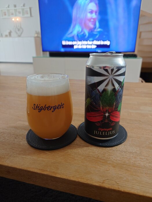 En ölburk och ett glas med Stigbergets Juleljus på ett träbord, med en tv i bakgrunden.