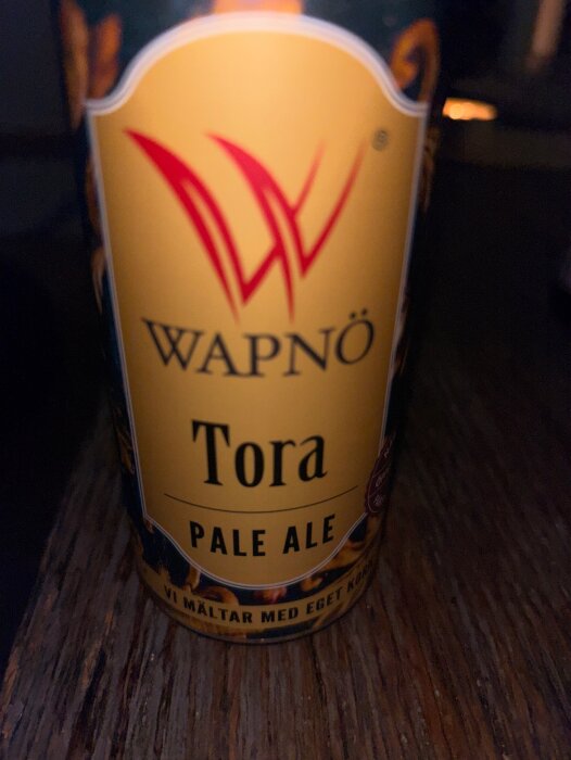 Wapnö Tora Pale Ale flaska, gul etikett med röd logotyp, mot en trämöblerad bakgrund.