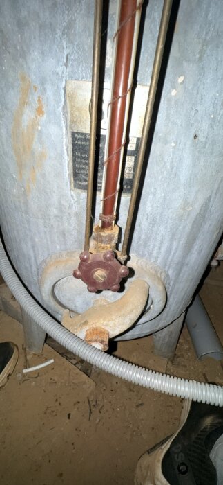 Järnrör med röd ventil kopplad till en trycktank, ansluten med grå plaströr på markyta.