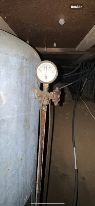 Manometer på en tank med rör och ratt, visar lågt tryck. Bakgrund med kablar i ett trångt utrymme.
