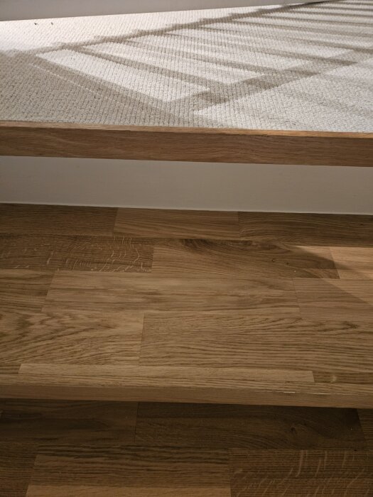 Trätrappa där sista steget är på plats. Mild skugga över stegen och en mattkant synlig på översta planet. Naturliga träådringar framträder tydligt.