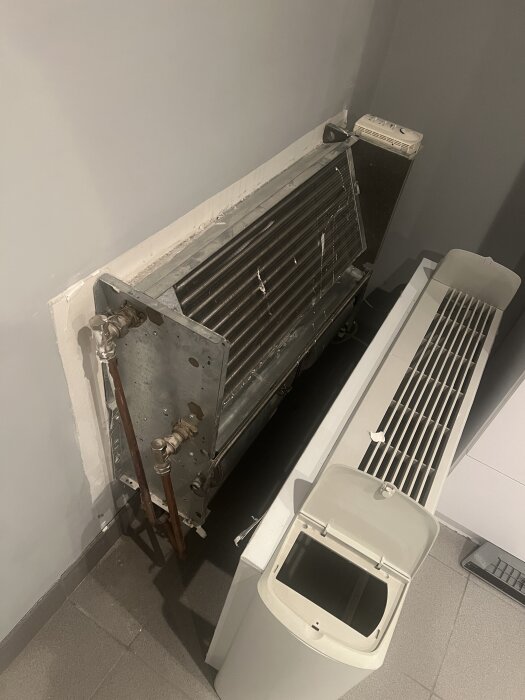 Fläktkonvektor monterad på vägg med exponerade rör och avtagbart hölje, sedd från sidan i ett rum.