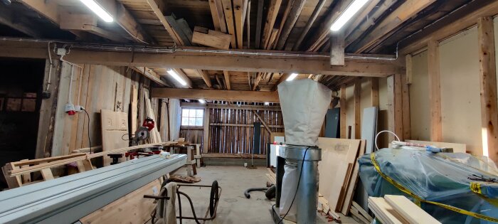 Snickeriverkstad med verktyg, träbjälkar i taket, plankor och material på golvet, och ett fönster som släpper in dagsljus.