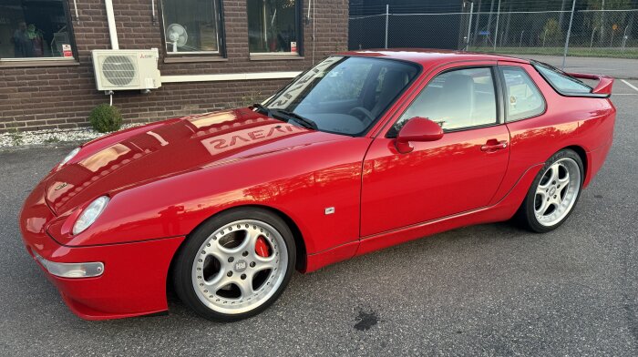 Röd sportbil med silverfälgar parkerad framför en tegelbyggnad.