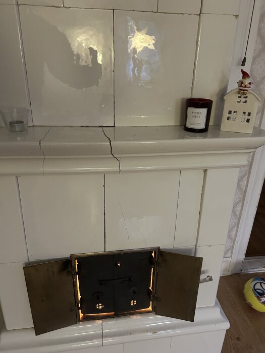 Spricka i vit kakelugn med öppna mässingsluckor, synliga dekor och ljus; fråga om reparation av sprickan vid eldning.