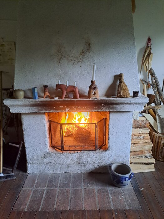 Öppen spis med brinnande eld, omgiven av prydnadsföremål på spiselkransen och ved staplad bredvid på ett trägolv.