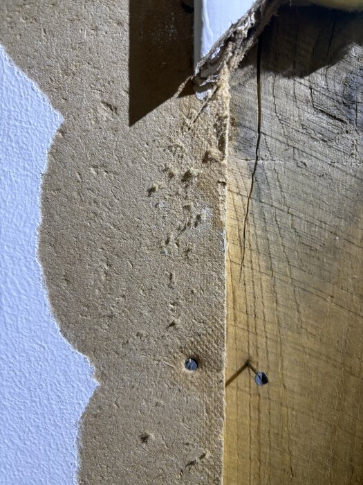 Närbild av en vägg med en uppfläkt sektion som visar brunt skivmaterial intill trä, från ett byggprojekt daterat 1957, med synliga spikhål.