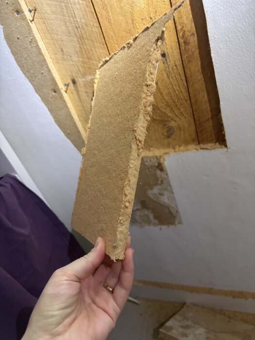 Hand håller en brun skiva från en vägg, misstänkt vara tretex eller masonit, med synligt trämaterial bakom från 1957.