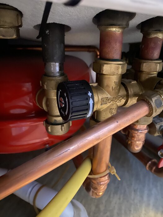 Bilden visar en närbild av rör och en blandningsventil med en justerbar ratt, inställd mot plussidan, på en värmepanna.