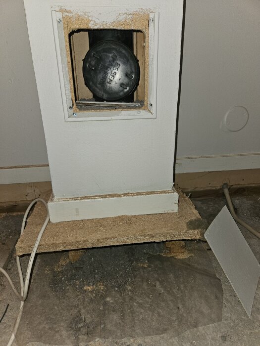 Svart plaströr med ESSEN-slangmuff bakom vit väggpanel, synlig genom öppning. Under panelen är det betonggolv, spånskiva och lösa kablar.