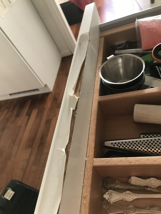 Öppen kökslåda med trasig kantlist och diverse köksredskap inuti, inklusive en metallskål och en osthyvel.