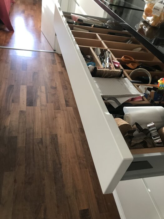 Öppen vit kökslåda med träinsats visar köksredskap, papper och snöre. Lådan verkar behöva en reparation eller utbyte.