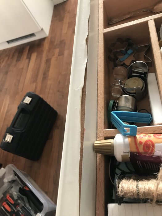 En kökslåda med skadat sidostycke och diverse föremål inuti, inklusive ett limtub och bakformar. Golvet och verktygslådor syns i bakgrunden.