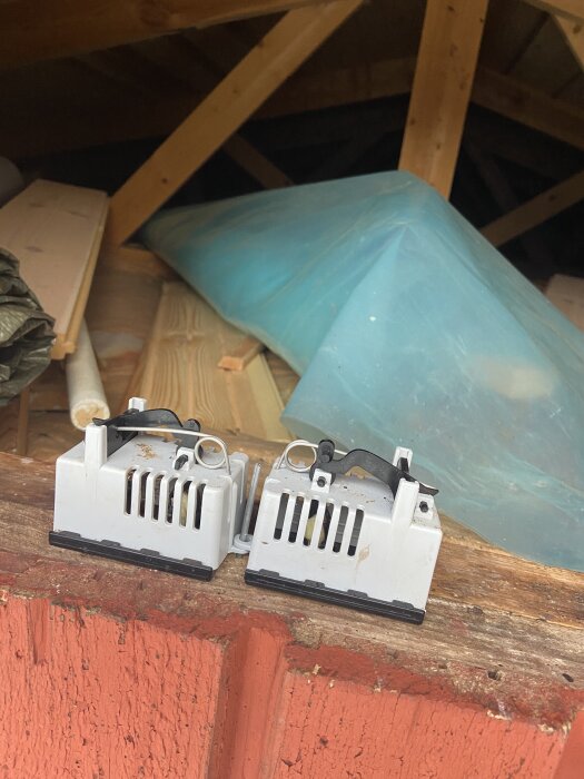 Två vita råttfällor står på en röd träbalk i vad som ser ut som en vind med träbjälkar och täckplast i bakgrunden.