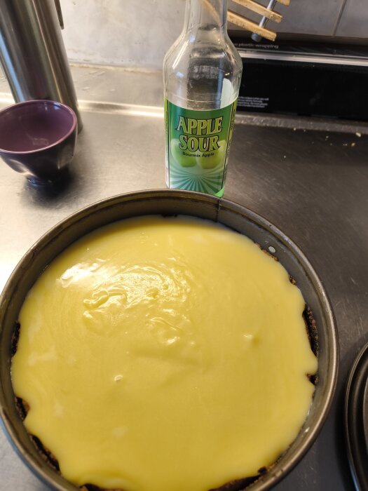 Cheesecake med gul topping i en form, bakgrund med flaska märkt "Apple Sour", lila skål och köksyta.