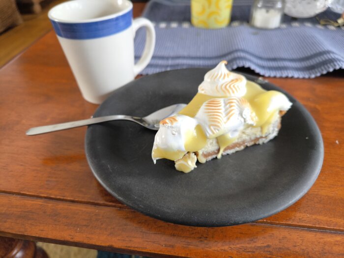Cheesecake med italiensk maräng och gul sås på svart tallrik, intill en mugg på ett träbord.