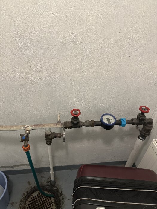 Vattenrör med två röda ventiler, en mätare och kopplingar, samt en blå och en orange slang kopplad till rörsystemet mot en vit vägg.