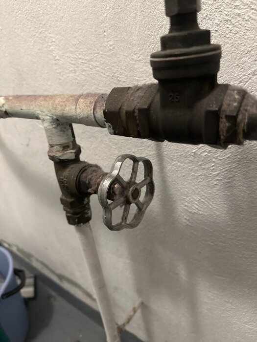 Gammalt vattenrör med ett rostigt, silverfärgat vred monterat mot en vit vägg, diskussion om vattenledningens funktion och placering.