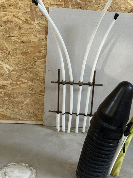 Vägg med gips- och OSB-skivor, vit rördragning och svart ventilationsrör på ett garagegolv med golvvärme.