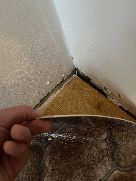 Detaljbild av en upplyft gammal plastmatta, med vit kärna, limmad på spånskiva. Mönster på mattan syns i nedre delen av bilden.