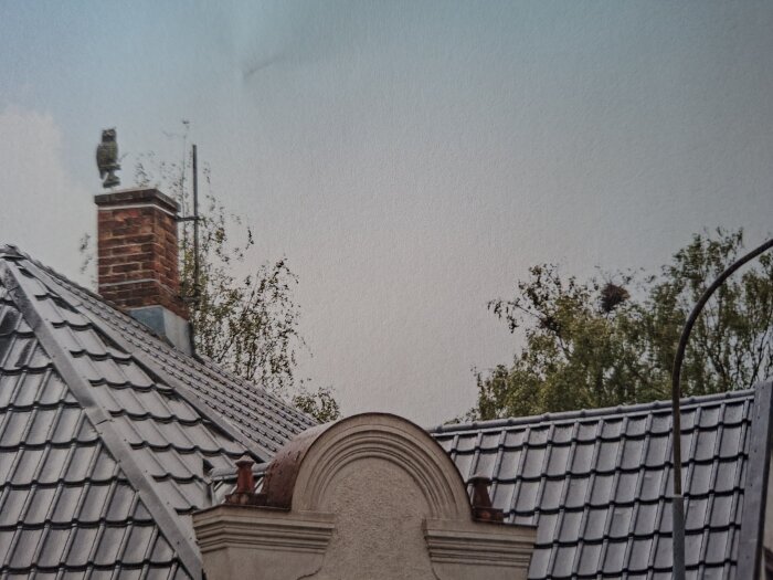 Tegelklädd skorsten med falsk uggla på ett tak med grå takpannor, omgiven av lövträd i bakgrunden.